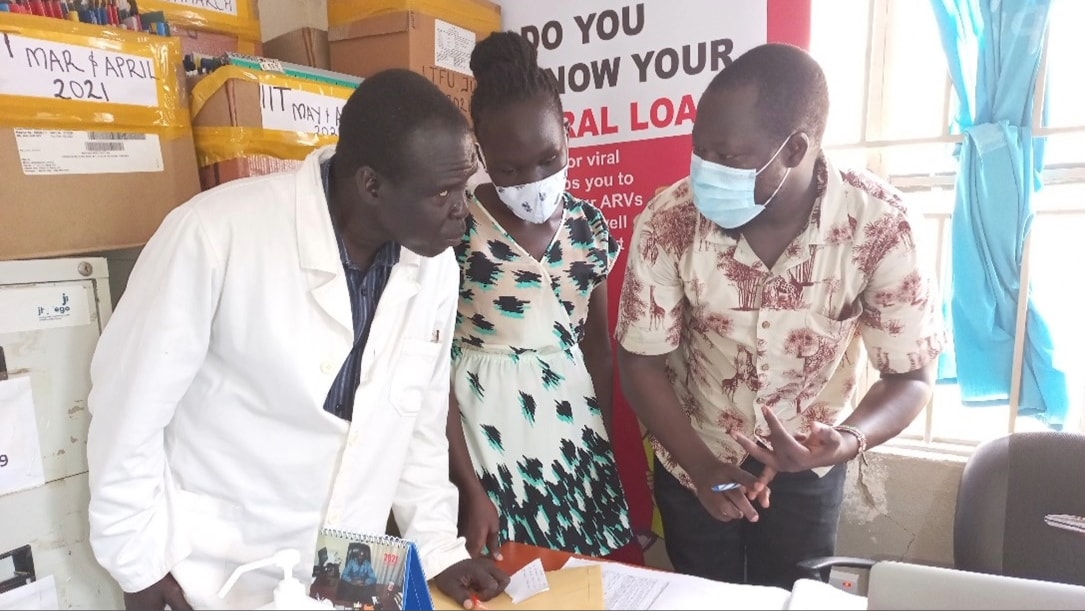 Three people stand behind a desk and talk. A poster behind them reads, "Do you know your viral load?"