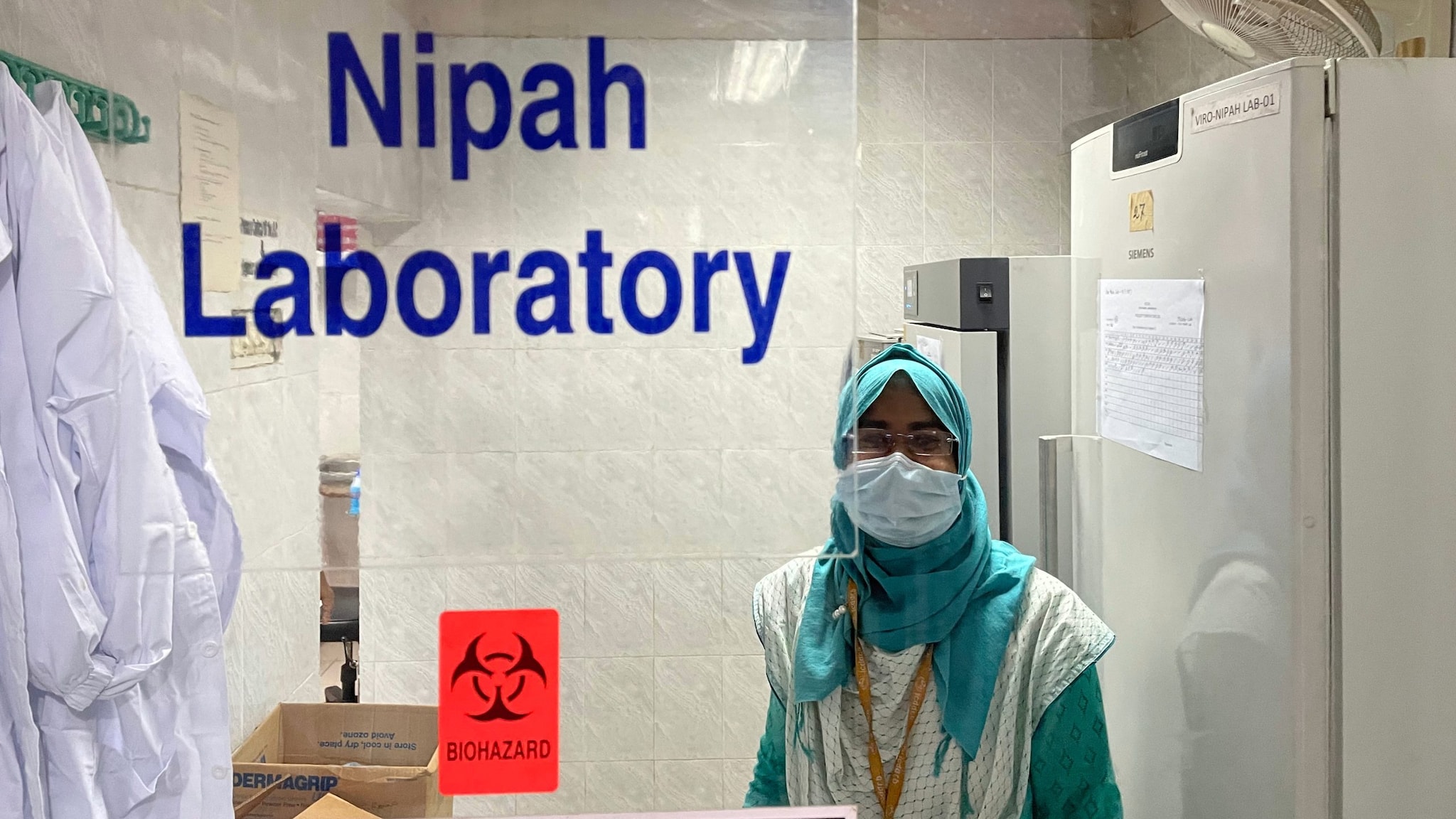 A person in a mask and head scarf stand behind a clear divider that has the words "Nipah Laboratory" and "Biohazard" on it.