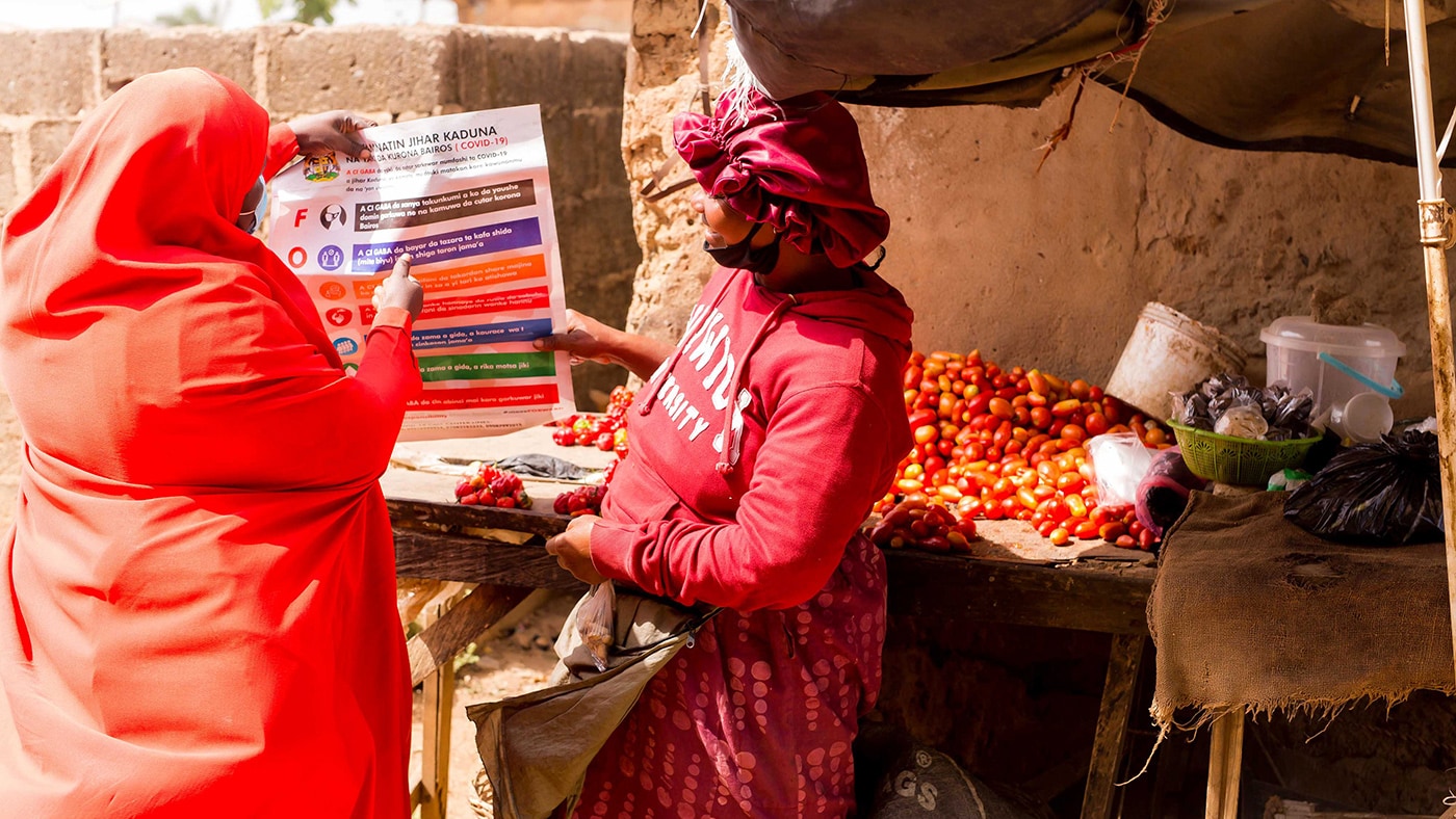 An individual in bright orange holds up an informational poster to show a woman who is wearing red as they stand outside.