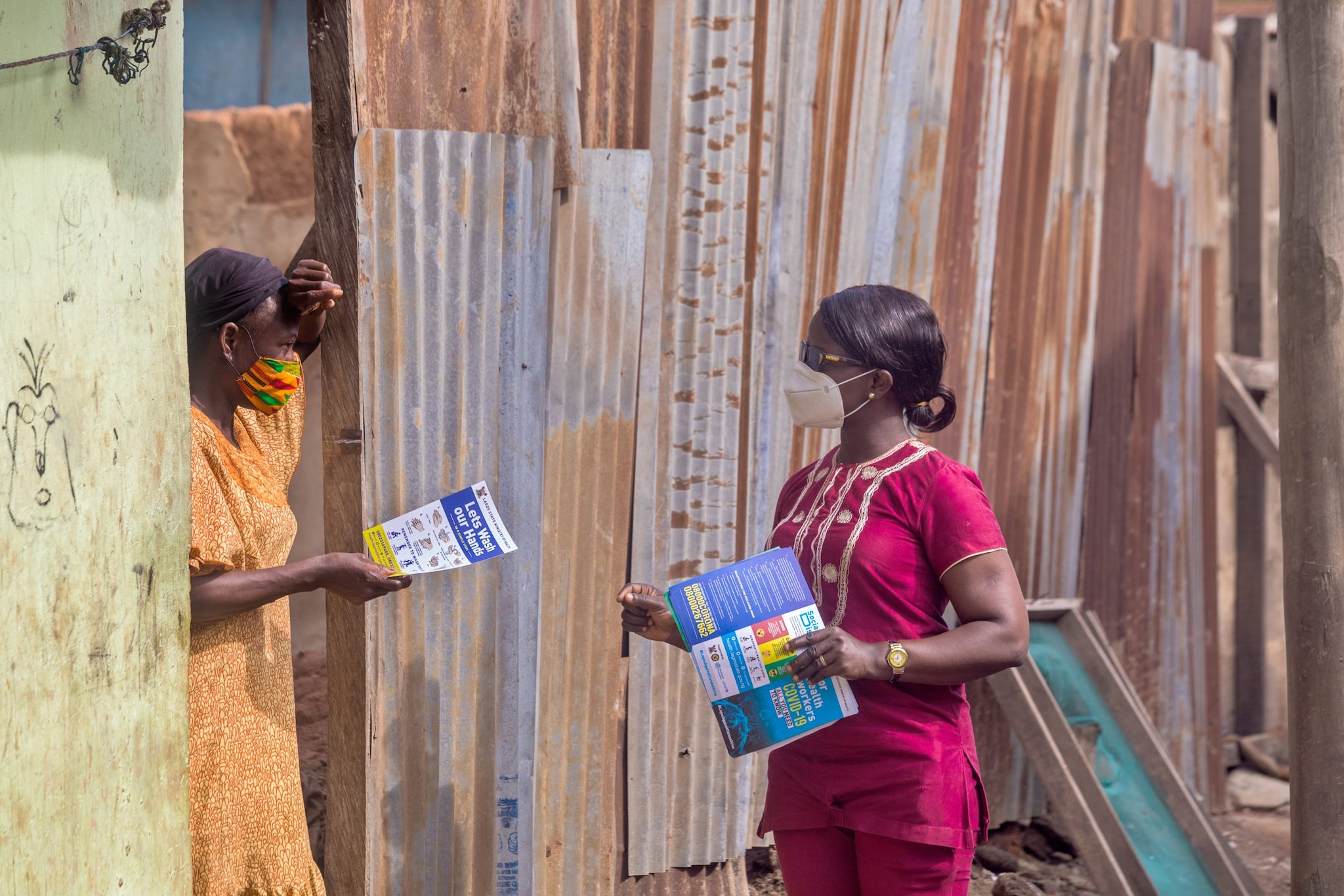 Two people talk outdoors while holding printed documents about handwashing and COVID-19 prevention.