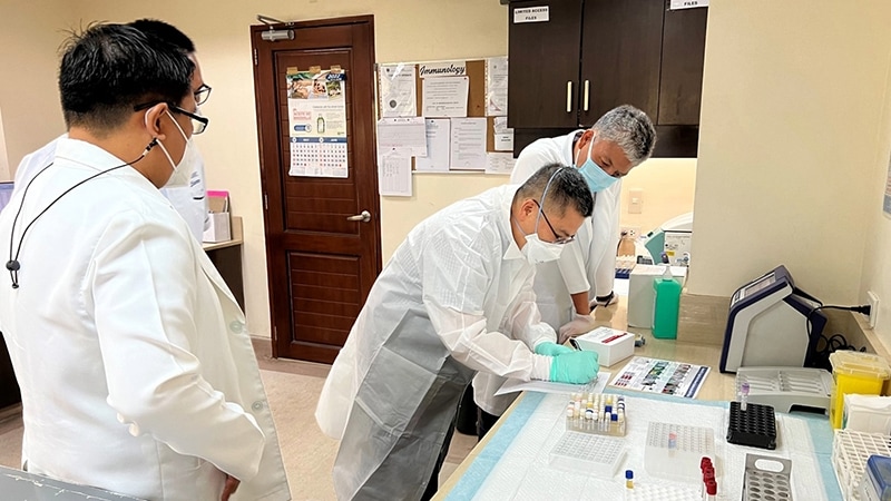 4 laboratorians stand in front of a desk with test tube trays on it. One person is writing down something while the other people look on.