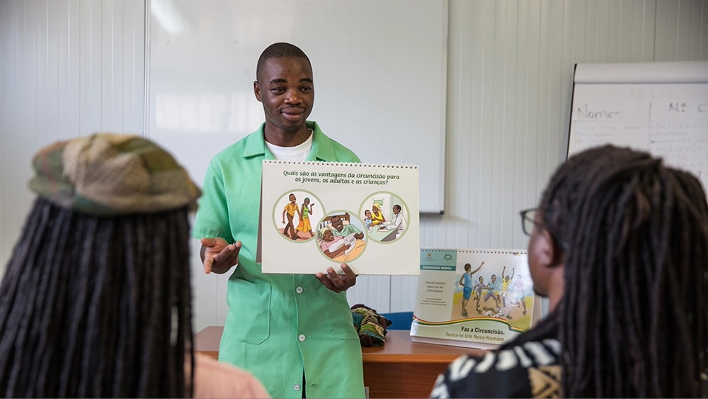 A health educator stands in front of two people and holds a poster