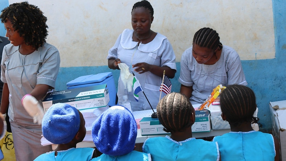 Four students in blue uniforms sit waiting for their vaccinations. Three health workers in pale blue dresses stand in front of the students.