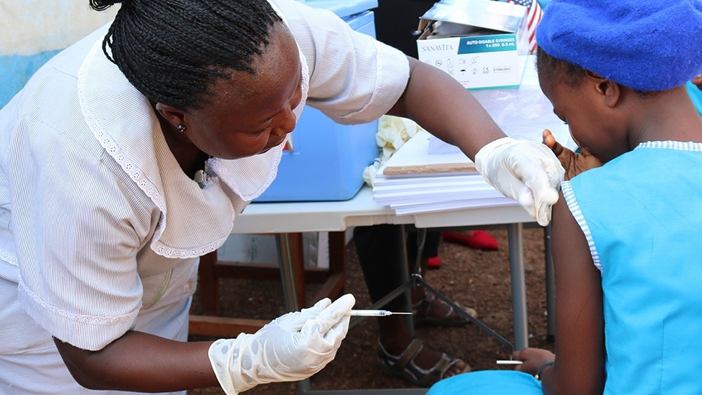 A health worker in a white outfit administers a vaccine to a young student who is wearing a blue school uniform.