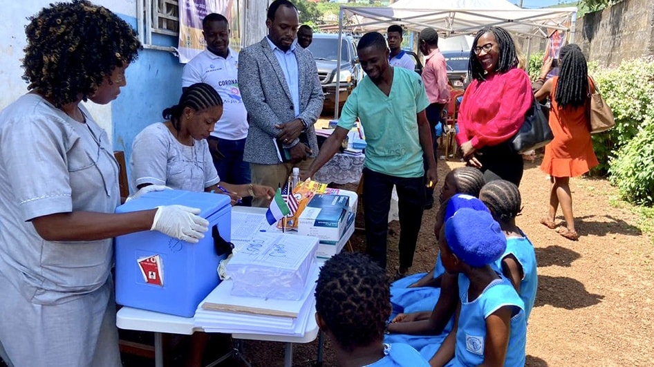 A group of health workers, health officials, and students gather outside a local school. Health workers are preparing to administer vaccines to the students.