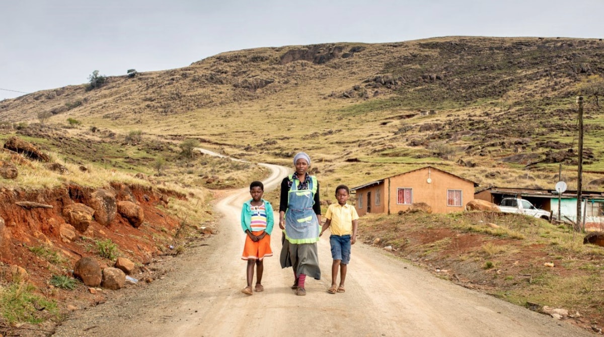 A woman with a young boy and girl beside her on road. Behind them is a house.