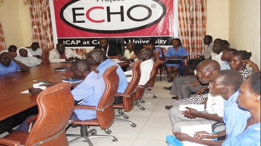 A group of people are seated around a table and along a wall. A sign behind them reads "Project ECHO: ICAP at Columbia University."