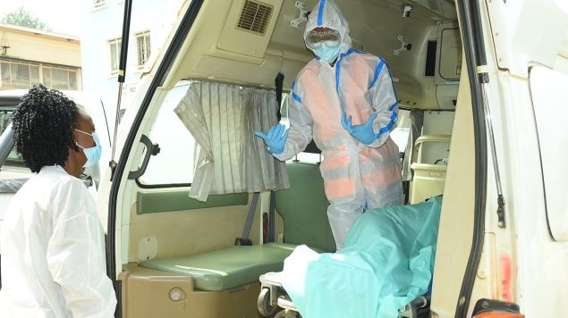 One health worker stands inside an ambulance as they talk to another health worker outside of the vehicle. Dressed in personal protective equipment, they prepare to transport a patient to a local hospital.