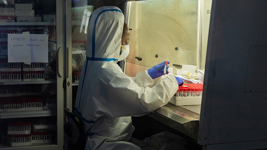 A lab technician in personal protective equipment sits and works with testing supplies.