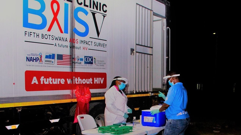 Three individuals in personal protective equipment stand in front of a BAIS V truck with coolers of medical samples.