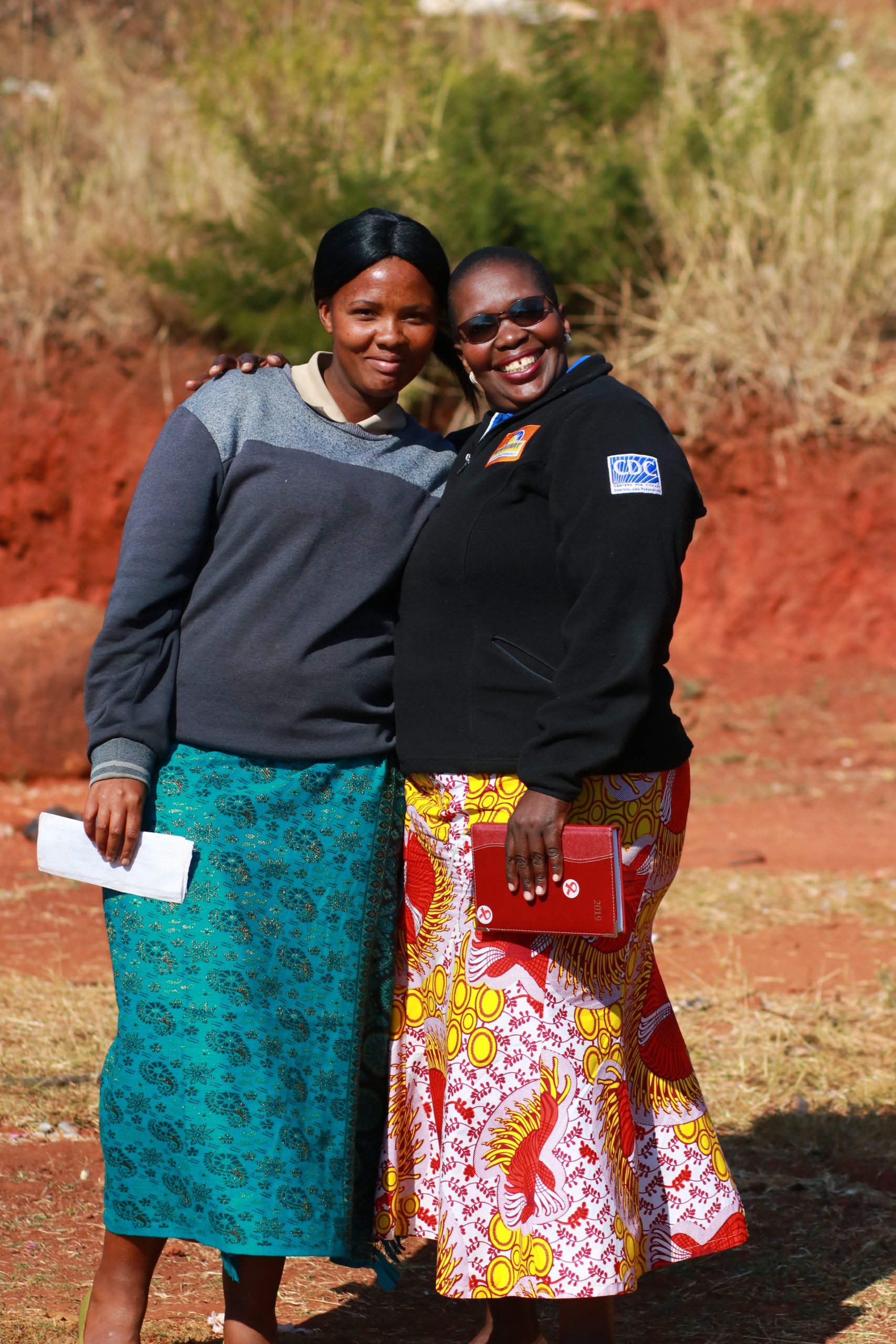 Two women stand on a dirt road smiling.