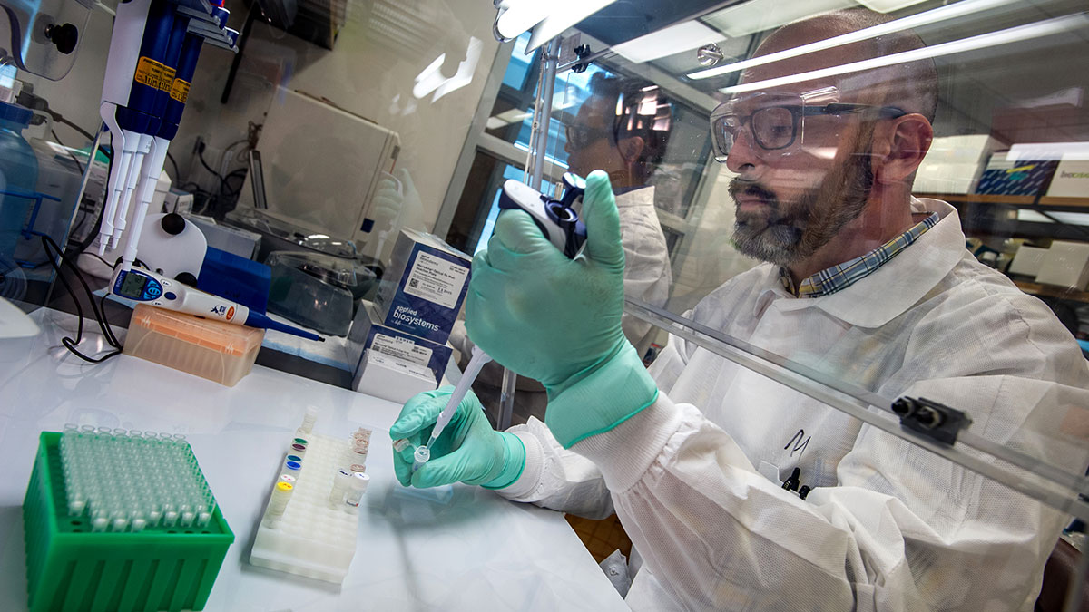 A man wearing a white coat, goggles, and gloves uses a pipette to transfer liquid into a test tube.