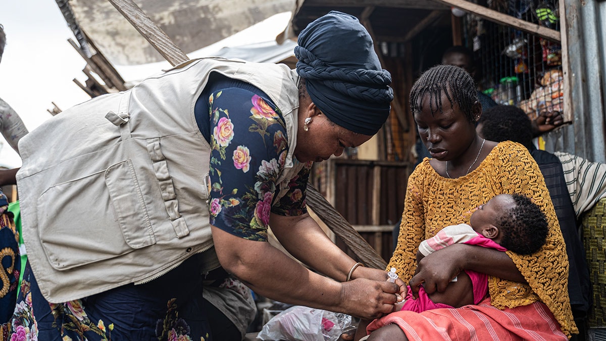 Outside of a wooden building, a woman wearing a khaki vest and a blue head scarf leans over another woman, who is holding an infant in her lap, and gives the infant a shot in her leg.