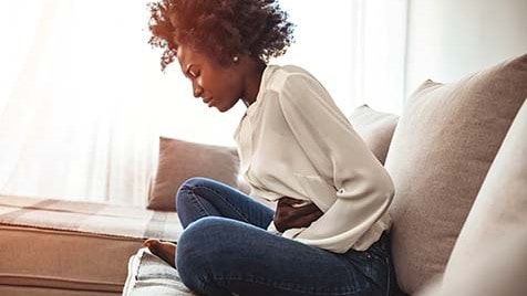 Woman sitting on a couch holding her stomach with her eyes closed and a pained look on her face