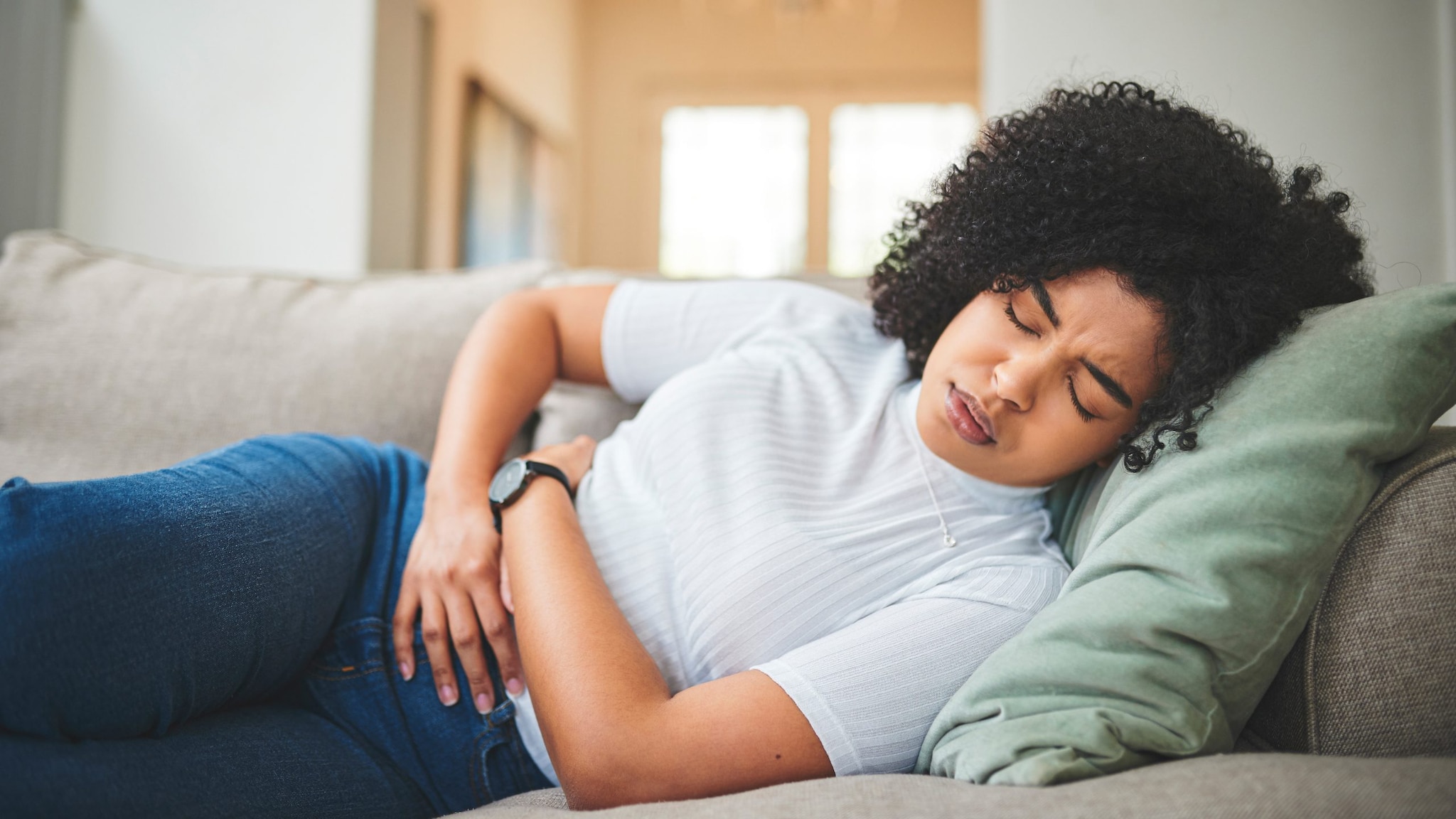 A young woman lays on a couch holding her stomach in pain