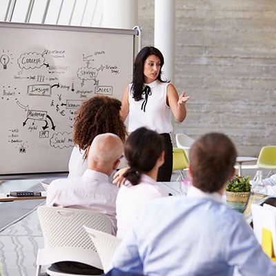 A woman in front of a white board speaking to a class of adults.