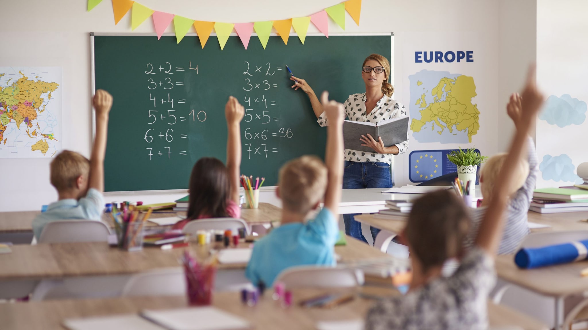 Teacher in front of class with engaged students raising their hands to answer question.