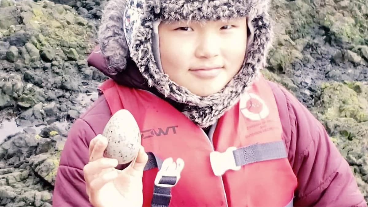 Native Alaskan boy in winter coat and life vest on rocky shore holding up a bird's egg.