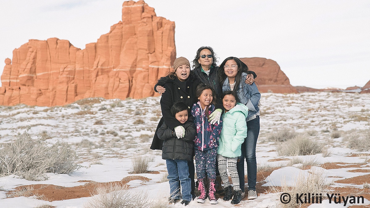 Older American Indian woman hugging two younger women and three younger children in front of them.