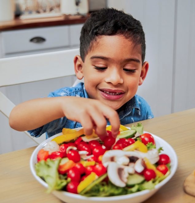 A little boy picking vegetables from a bowl on a kitchen counter.