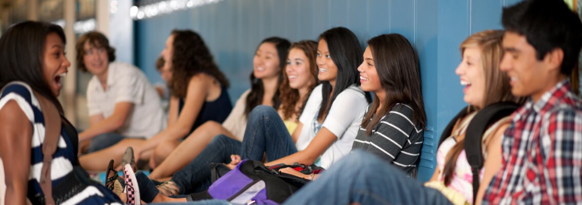 Large group of diverse youths conversing while sitting at the foot of a line of lockers