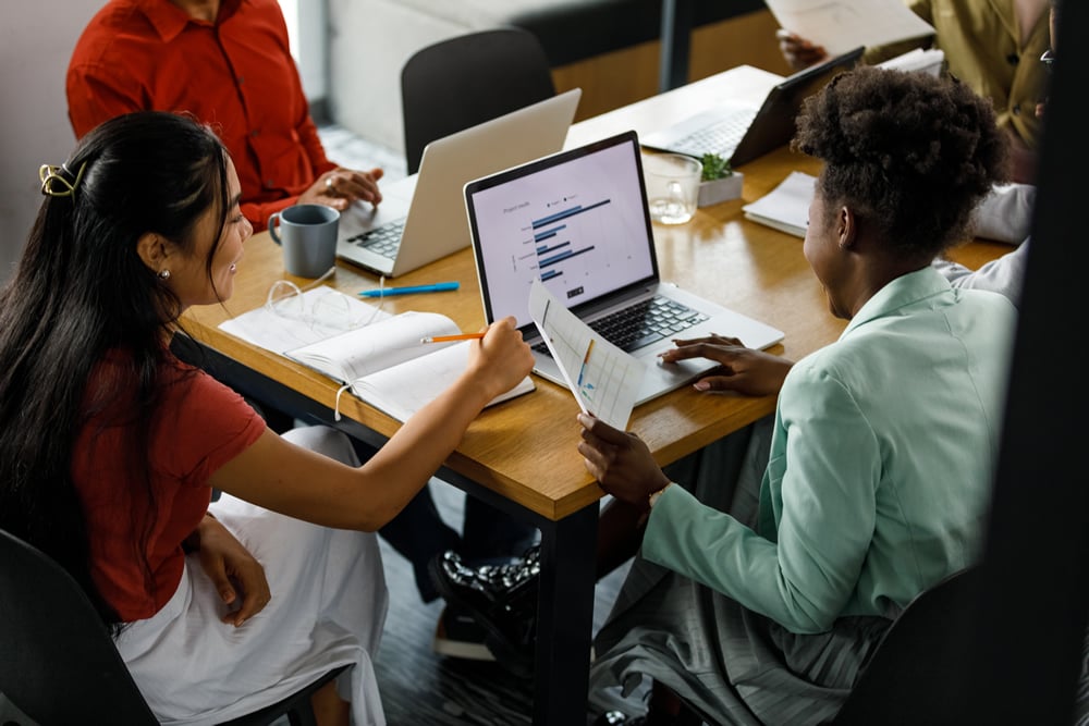 View of two young female students reviewing a document and data on laptop with members during a youth advisory meeting.