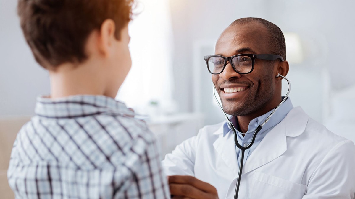 Black pediatrician in a white lab coat using a stethoscope to listen to a child’s heart.