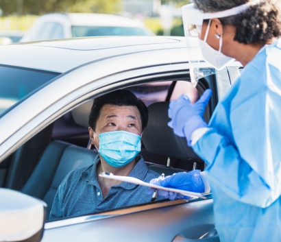 Masked person sitting inside a car talking to another person with PPE on standing outside at a COVID-19 testing site.