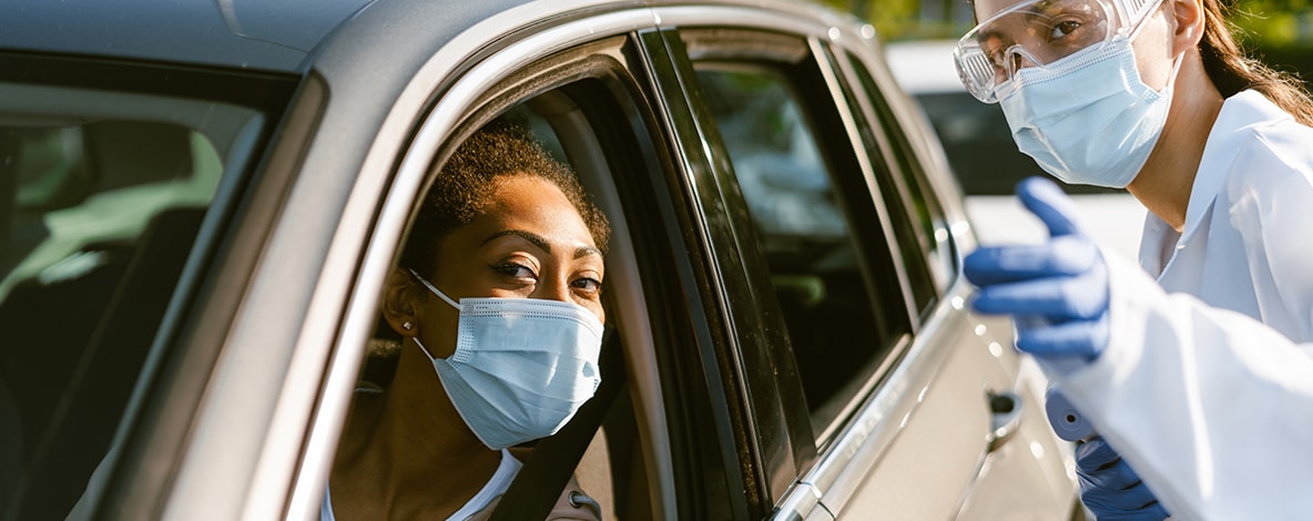 Masked person in car getting instructions from another person with PPE on outside of the car at a COVID-19 testing site.