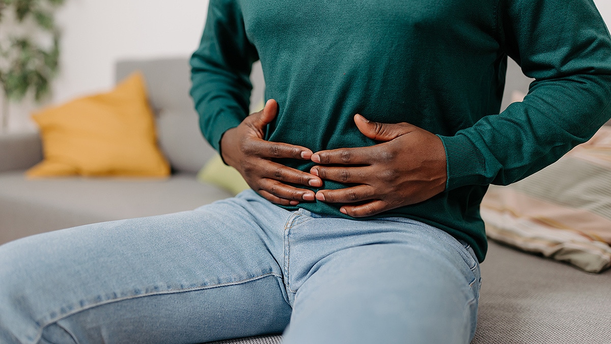 A young man sits on a sofa, holding his stomach from digestive pain.