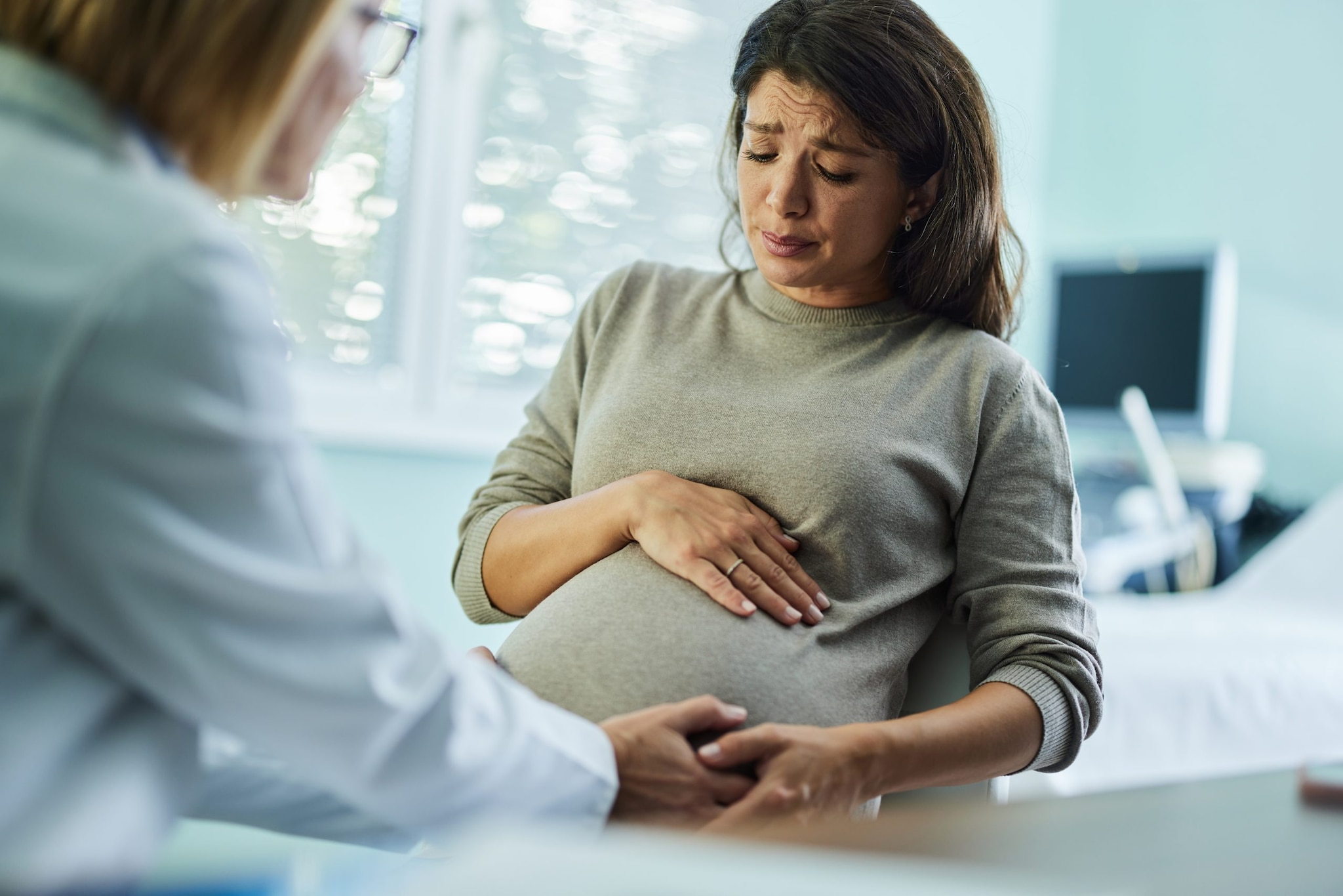 A pregnant person holding their pregnant stomach and looking worried while speaking to a doctor in a medical office.