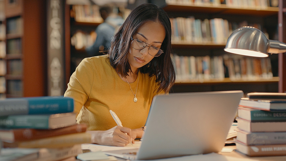 Dark-haired woman in a library with laptop doing research.