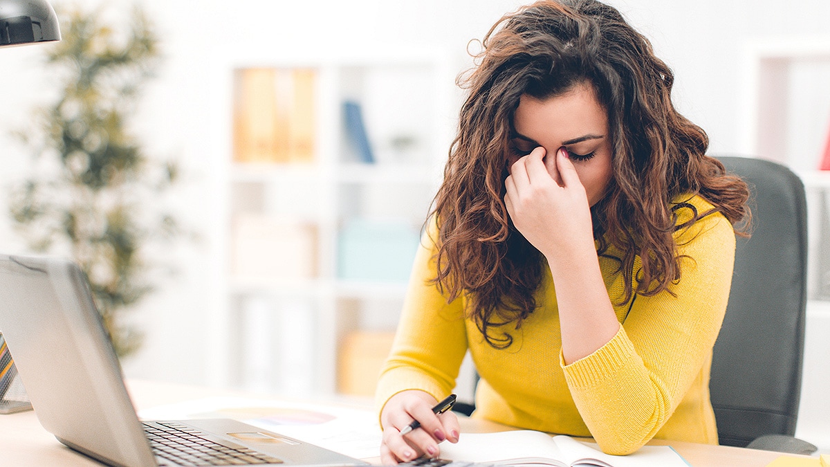 Young woman at desk with hand to bridge of nose and eyes closed.