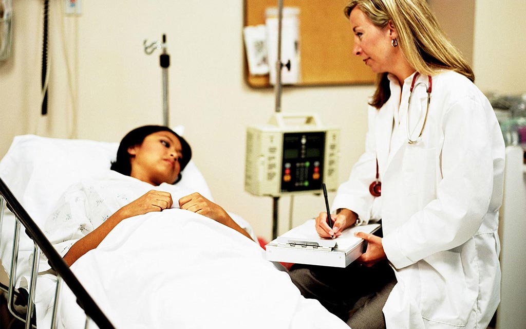 A female patient lying in bed at a hospital or clinic, with a female doctor sitting by her bedside with w clip board.