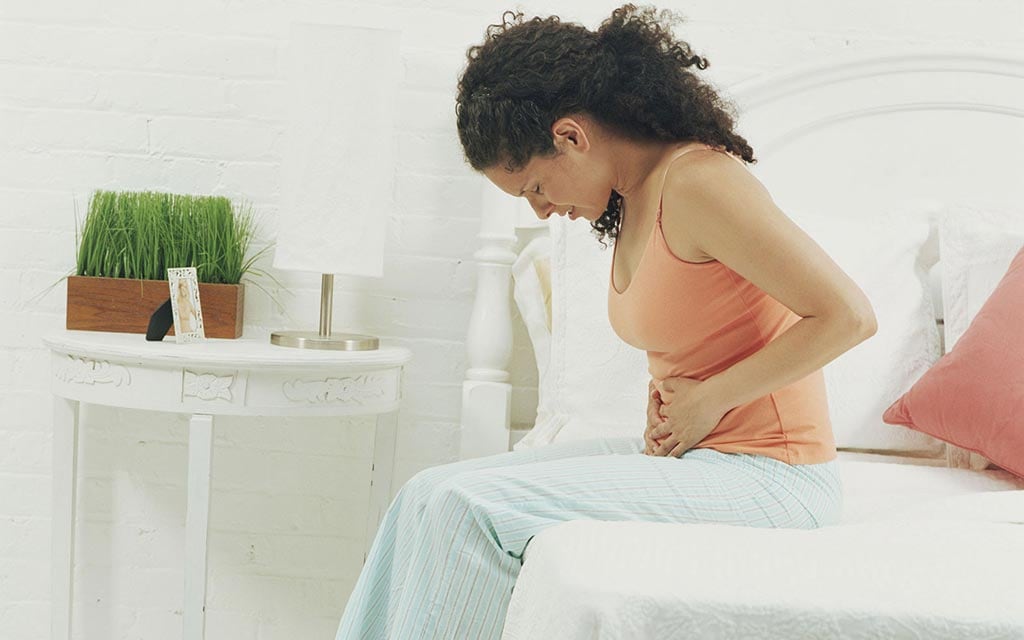 A young woman sitting on a bed holding her with stomach.