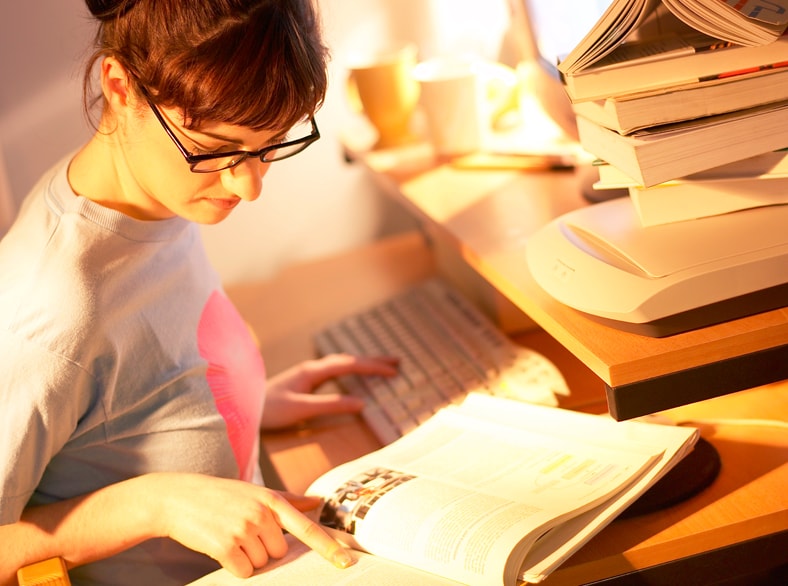 Young woman reading through books at a computer.