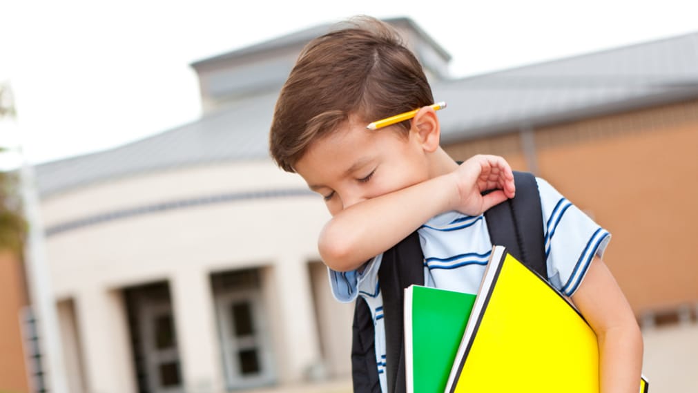 A young boy covers his cough in his elbow while leaving school.