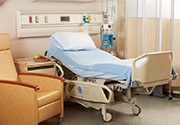 Empty hospital bed in a room with a chair, table, and medical equipment next to it.