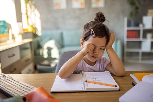 Tired young girl rubs her eyes as she sits at a table trying to complete her homework.