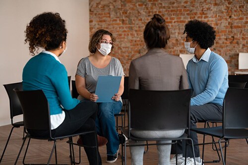 People of various races wearing masks sit in a circle talking.