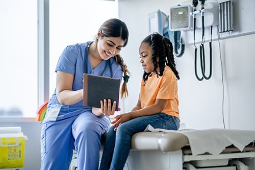 A young girl sits on an exam table during a routine checkup. The girl smiles gently as her healthcare provide reviews medical information with her on a tablet.