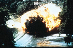 A giant fireball surrounded by Black smoke, followed by red fire and then white fire coming from the direction of several 2 or 3 story buildings nestled into trees.