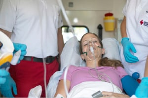 A young woman with long brown hair lies on a gurney in an ambulance. Her eyes are closed and she is wearing an oxygen mask and a blood pressure finger monitor. To either side of her, attending her, are a man and a woman whose faces are out of the picture frame. The woman is wearing a white uniform with a hospital cross and a pair of blue single-use gloves. The man is wearing a white polo, red pants, and blue single-use gloves.