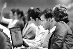 A row of men and women sitting in what looks to be an auditorium or large classroom. One student in the background is raising her hand. A young man and a graying woman are looking at a laptop computer together.