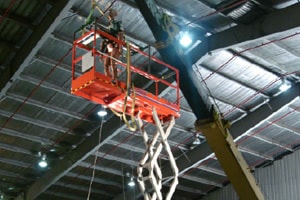 Inside an industrial space is a hydraulic lift with a red platform about 3 feet wide and 7 feet long. The platform is in the center of the picture. It is fully extended and there is a test dummy towards the front of the platform. There also appears to be the arm of a crane behind the lift.