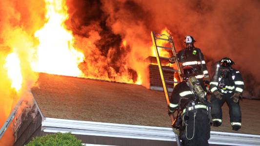 Three firefighters wearing protective gear working to extinguish a fire on the roof of a burning house. Firefighting concept