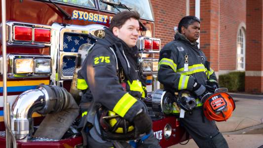 Two firefighters wearing protective gear, though with their helmets off, lean against a parked firetruck during a moment of rest. Firefighting, comradery concepts