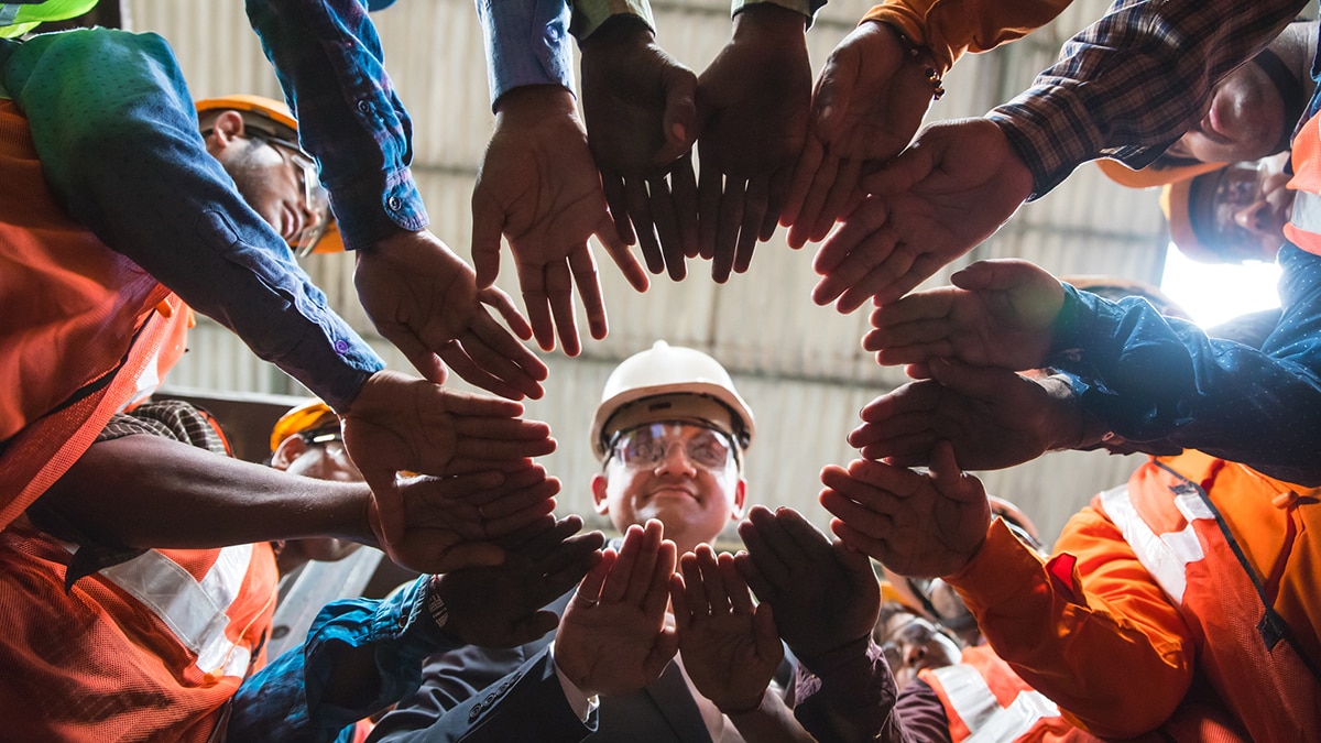 Owner looking down towards the camera in the center, with all his factory employees huddled up in a circle with their arms in the center.