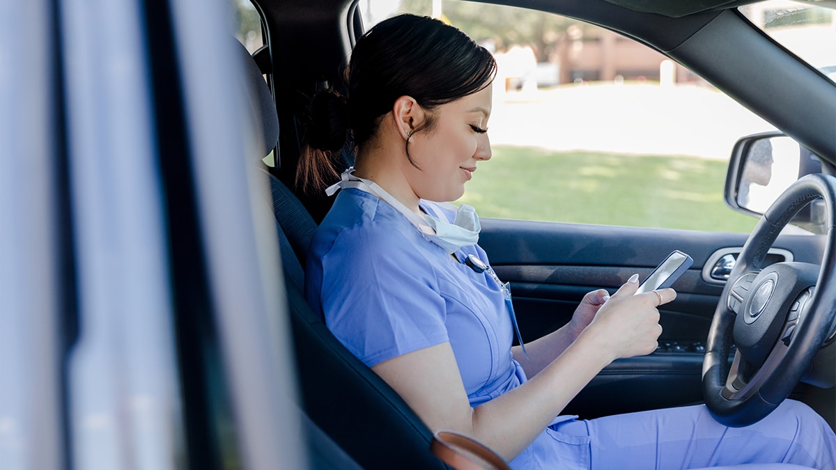 Healthcare worker in scrubs looks at her phone while sitting in the driver's seat of a parked car.