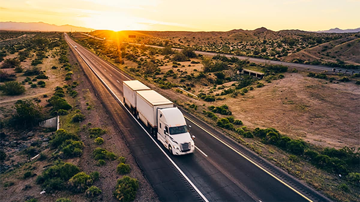 Photo of large white tractor trailer driving on a vast straight interstate indicative of the Western United States.